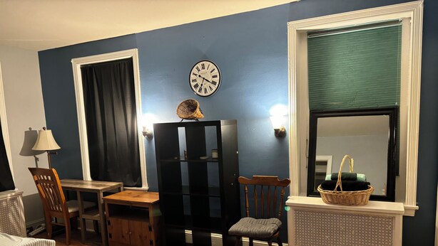 A room with a wooden floor, a black entertainment center, a wooden chair, a small table, a lamp, a clock, a window with a green blind, and a mirror.