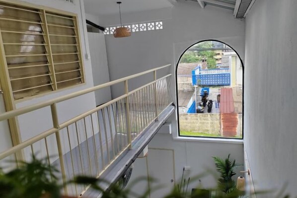 Un pasillo con barandilla metálica, una ventana con vista a un edificio azul, y una lámpara colgante.