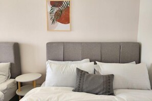 A hotel room with a grey upholstered headboard, a round wooden side table, and a framed artwork on the wall.