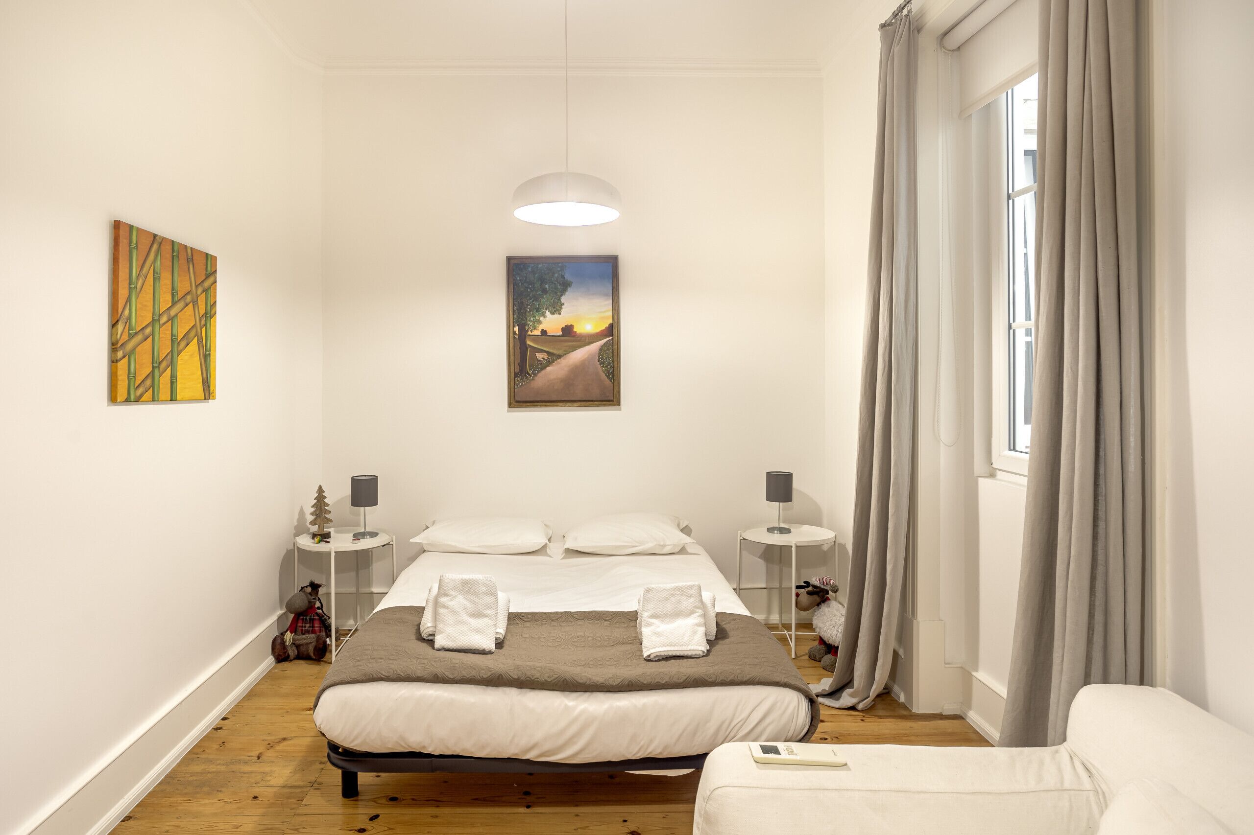 Un dormitorio con una cama, dos mesitas de noche, una lámpara, una ventana con cortinas y obras de arte en las paredes.