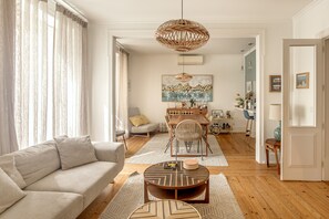 A modern living room with a sofa, coffee table, dining table, and chairs. There is a painting on the wall and a wicker chandelier. - Grand Central Apartment by Homing (Lisbon)