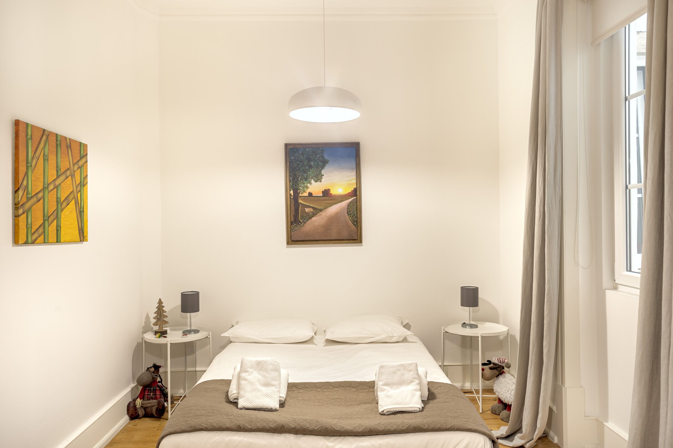 Una camera d'albergo con un letto, due comodini, una lampada, un quadro e una finestra con tende.