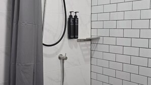 A shower area with a white tiled wall, a showerhead, and two bottles of shampoo on a shelf. - J.K. Mansion (Bangkok)