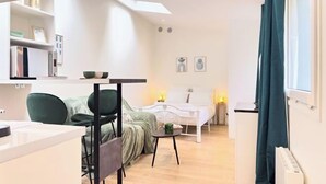 A modern, well-lit room with a bed, a small table, and a chair. There is a window with curtains, a radiator, and a shelf with books and decorative items. - Montparnasse 2 - Studio (Paris)