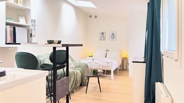 Ein modernes, helles Zimmer mit einem Bett, einem kleinen Tisch und einem Stuhl. Es gibt ein Fenster mit Vorhängen, eine Heizung und ein Regal mit Büchern und Dekoration.