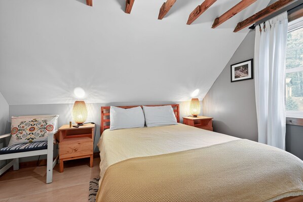 A bedroom with a sloped ceiling, a bed with a beige quilt, two bedside lamps, a wooden nightstand, a chair with a patterned cushion, and a window with curtains.