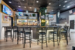 Zona de bar con varios taburetes, una barra y una pantalla de televisión que muestra un noticiero.
