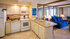 Una cocina compacta con microondas, estufa y lavavajillas, un pequeño comedor con mesa y sillas, y un sofá azul en una sala de estar con ventanas amplias.
