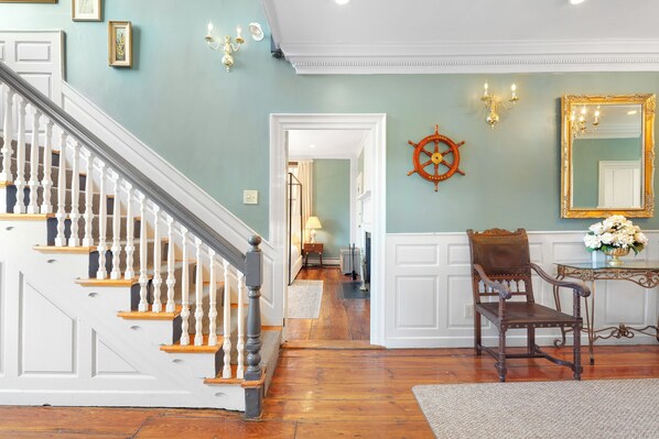 Un escalier avec des balustrades blanches et des marches en bois, une chaise en bois, un miroir avec un cadre doré et un gouvernail de bateau fixé au mur.