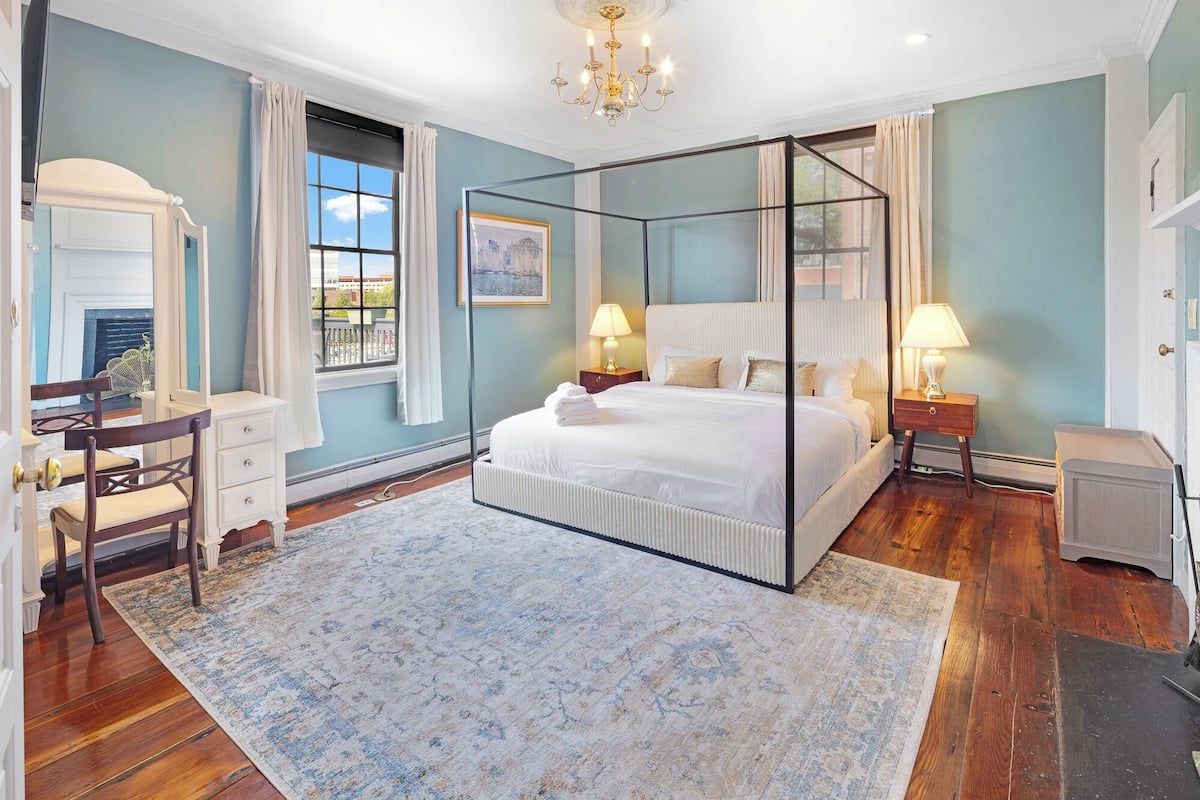 A bedroom with a four-poster bed, a sitting area with a chair and a table, and a view of the city through the window.