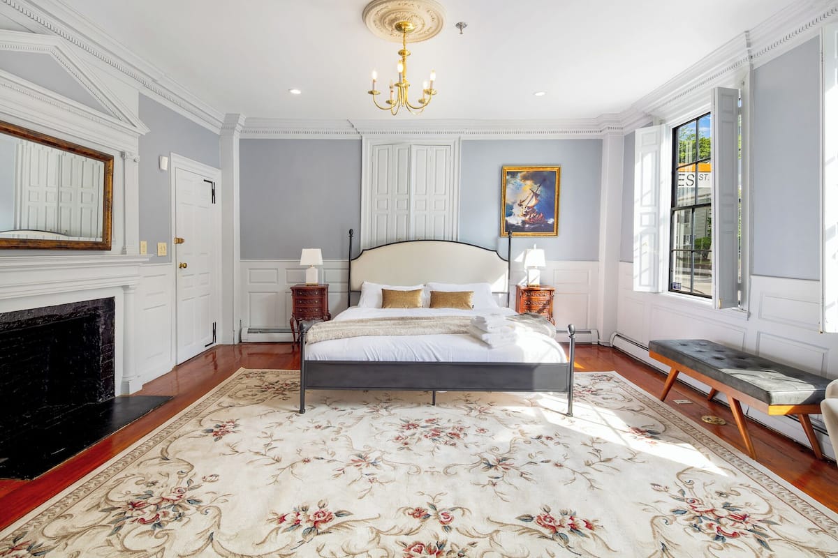 A spacious bedroom with a large bed, a fireplace, a chandelier, and a view of the street through a large window.