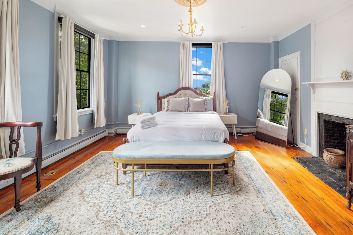 A bedroom with a large bed, a nightstand, a chair, a mirror, and a fireplace.