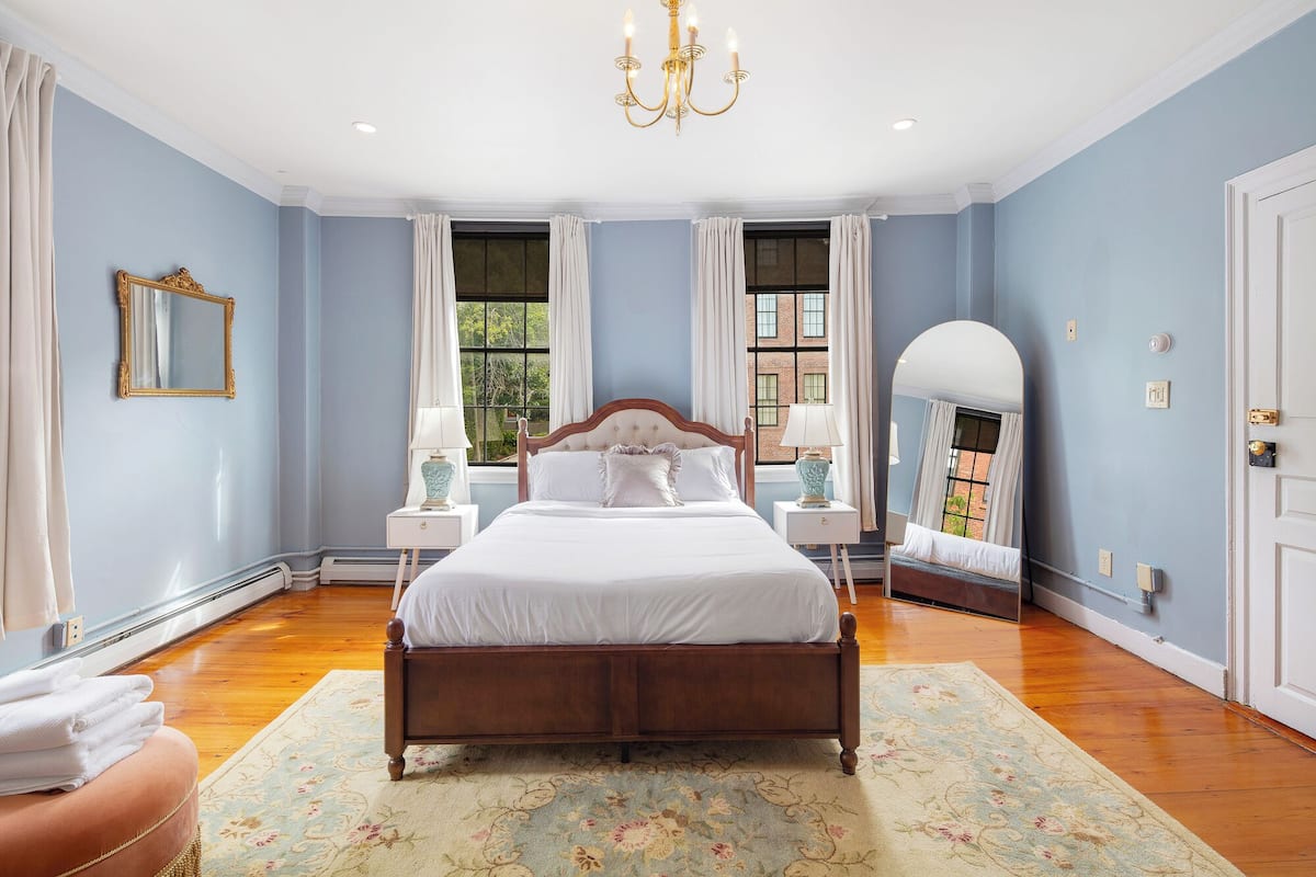 A bedroom with a large bed, two bedside tables, a chandelier, and a view of a brick building through the windows.