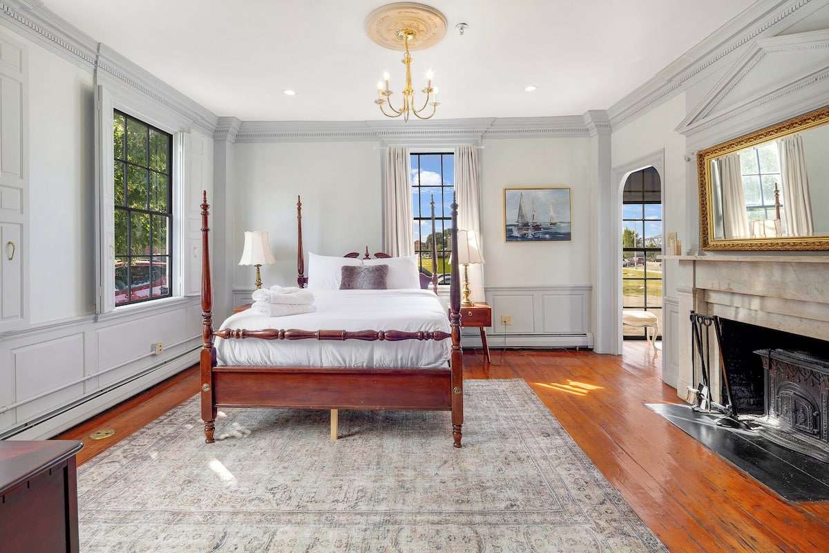 A spacious bedroom with a four-poster bed, a fireplace, and large windows offering a view of the outdoors.
