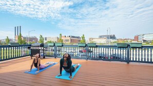 Two individuals on blue yoga mats on a wooden deck with a railing, overlooking a cityscape with buildings and a bridge. - The Hill (Providence)