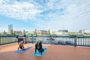 Dos personas sobre esterillas de yoga azules en una terraza de madera con barandilla, con vista a un paisaje urbano que incluye edificios y un puente.
