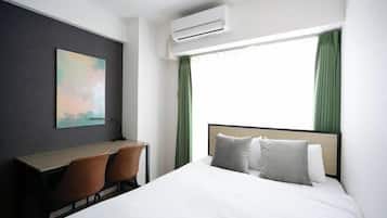 A hotel room with a bed, a desk, two chairs, an air conditioner, and a window with green curtains.
