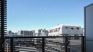 A view from a balcony with horizontal blinds, overlooking a cityscape with various buildings. - Daffitto Mitaka (Mitaka)