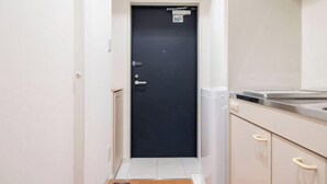 A narrow hallway leading to a dark door with a small sign above it, a white tiled floor, and a kitchenette area with a sink and cabinets. - Daffitto Mitaka (Mitaka)