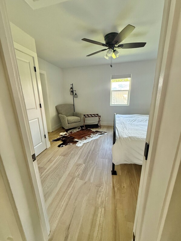 Un dormitorio con cama, una silla, ventilador de techo, ventana y piso de madera.