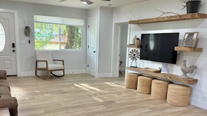 A spacious living room with a wooden floor, a ceiling fan, a TV mounted on the wall, and a window with a view of greenery outside. - 100 year old Montana Cottage! (Manhattan)