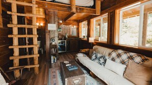 A cabin interior with a loft bed, wooden walls, a sofa with plaid cushions, a small table, and a window with a view of trees.