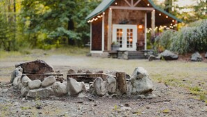A rustic cabin with a stone fire pit in front, surrounded by greenery.