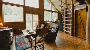 A rustic cabin interior with large windows, a wooden ladder, a dining area, and a cozy seating space.