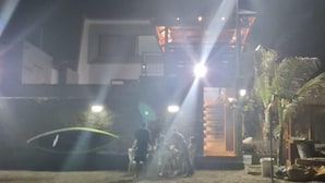 Cena noturna mostrando um grupo de pessoas reunidas em frente a um edifício, possivelmente um hotel ou restaurante, com luzes brilhantes e uma entrada bem iluminada.