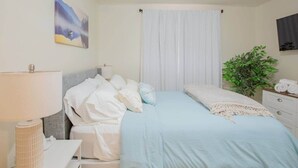 Un dormitorio con cama, mesita de noche, lámpara, planta y un cuadro en la pared.