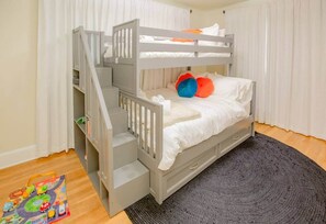Un lit superposé avec une structure grise, une literie blanche et des coussins colorés. Un petit escalier mène au lit supérieur, et un tapis gris recouvre le sol.