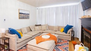 Ein Wohnzimmer mit einem beigefarbenen Ecksofa, einem kleinen Tisch mit Lampe, einem Kamin und einem großen Fenster mit weißen Vorhängen.
