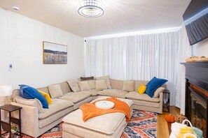 A living room with a beige sectional sofa, a small table with a lamp, a fireplace, and a large window with white curtains.