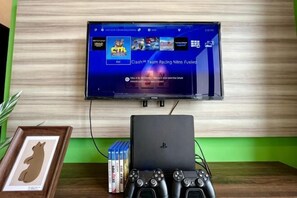 A flat-screen TV mounted on a wall, a PlayStation console, and a gaming controller on a table. - RiverView 8Pax 3Br in KBtown PS4, Netflix A-1-2 (Kota Bharu)