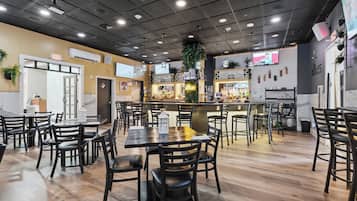 A restaurant interior with multiple dining tables and chairs, a bar area, and a television screen mounted on the wall.