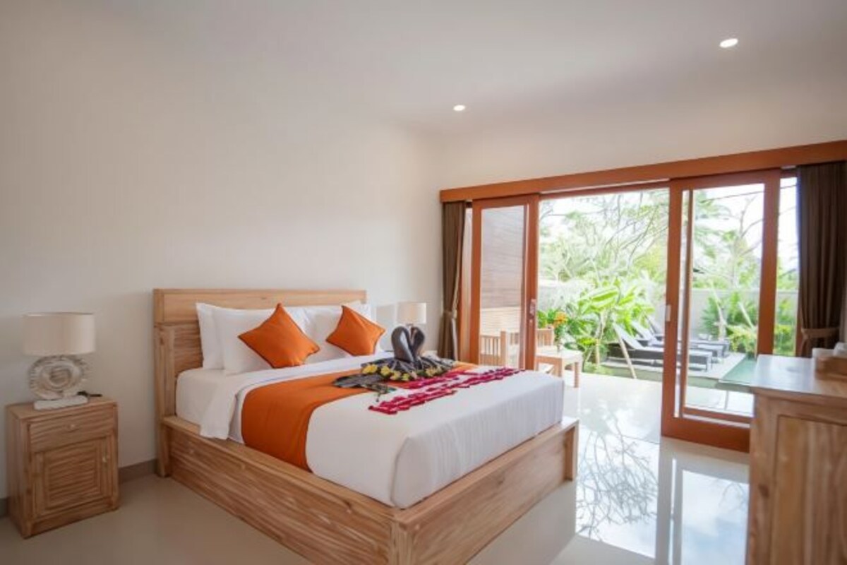A bedroom with a wooden bed, a nightstand, a desk, and a view of greenery outside.