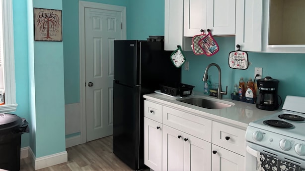 Una cocina con paredes verde azulado, armarios blancos, un refrigerador negro, una estufa y un lavamanos. Hay dos toallas de cocina colgadas y una cita enmarcada en la pared.