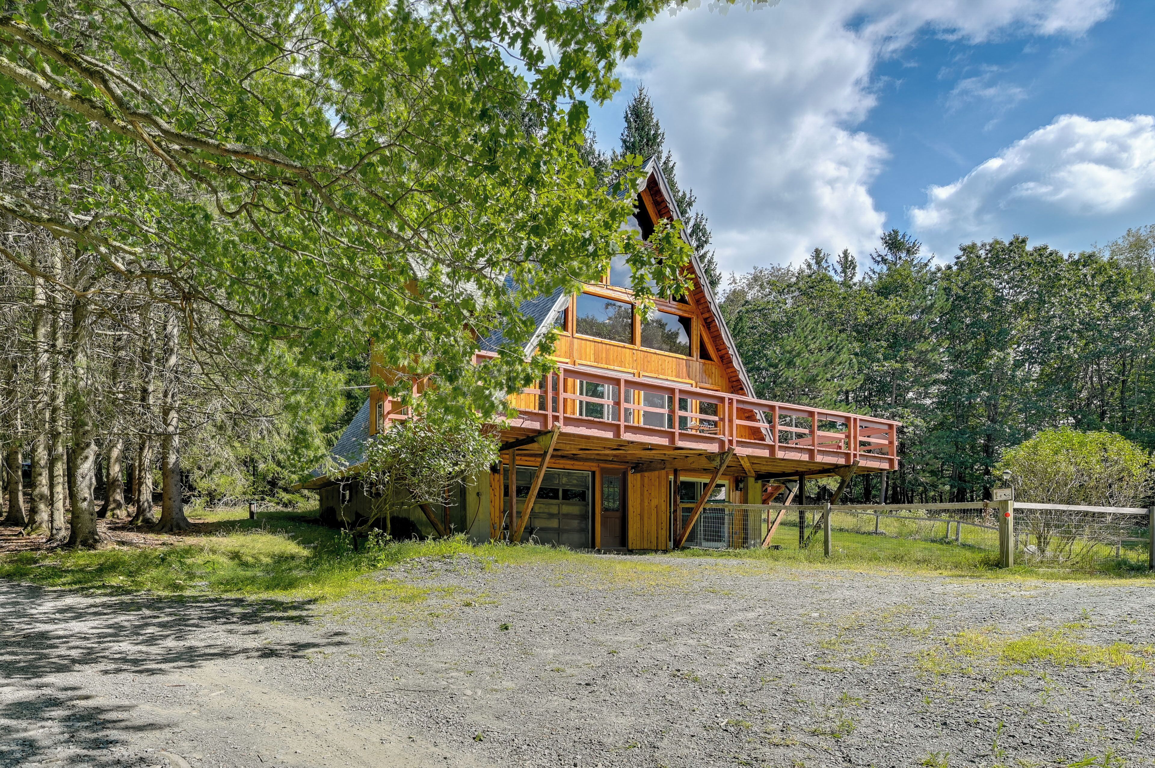A rustic cabin with a wooden deck and large windows, surrounded by trees and a gravel driveway.