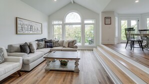 Een ruime woonkamer met een hoge plafond, een groot raam, een zithoek en een eethoek met stoelen.