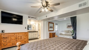 Quarto com cama grande, cômoda de madeira, ventilador de teto e televisão na parede.