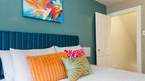 Een slaapkamer met een blauwgroen hoofdeinde, wit beddengoed en kleurrijke kussens.