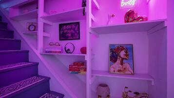 A staircase with white railings and shelves containing books, a neon sign, and a framed painting.