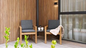 Ein gemütlicher Sitzbereich mit zwei Holzstühlen und einem kleinen Tisch, ein Fenster mit Vorhängen und einer Holzwand.