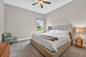 A bedroom with a large bed, a ceiling fan, a window, a chair, and a bedside table with a lamp.