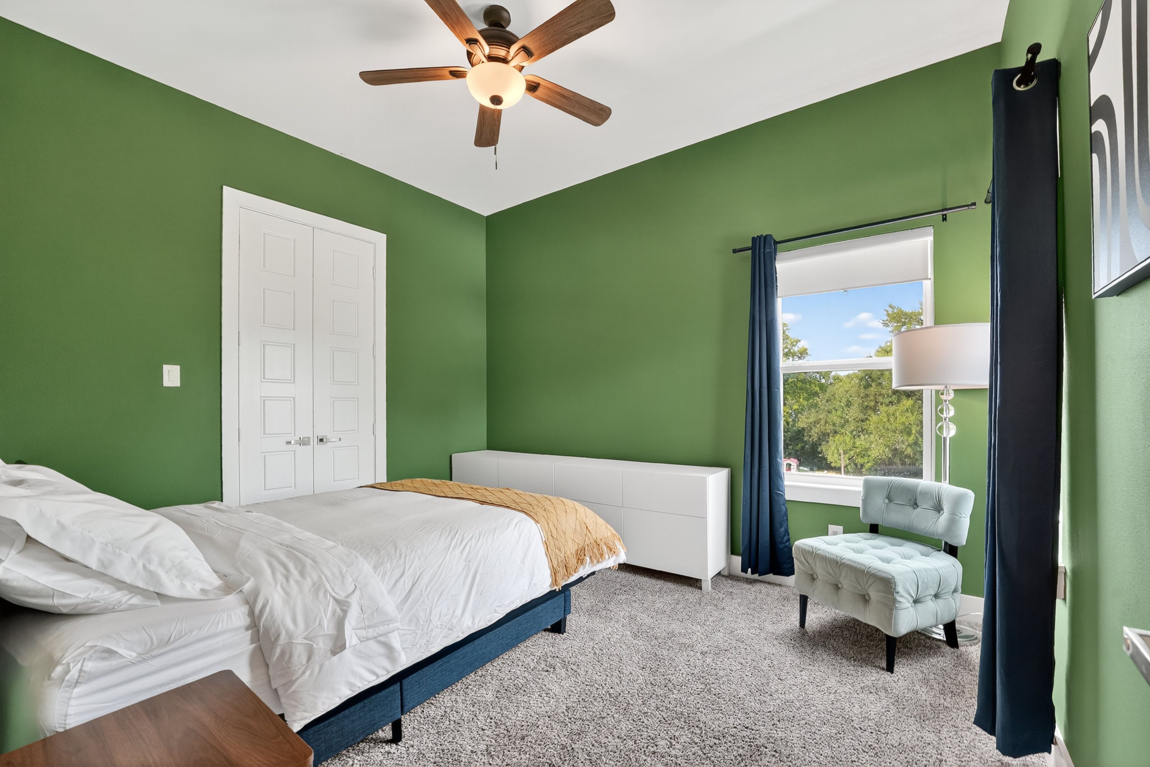 A bedroom with a bed, a chair, a window with curtains, a ceiling fan, and a door.