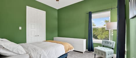 A bedroom with a bed, a chair, a window with curtains, a ceiling fan, and a door.