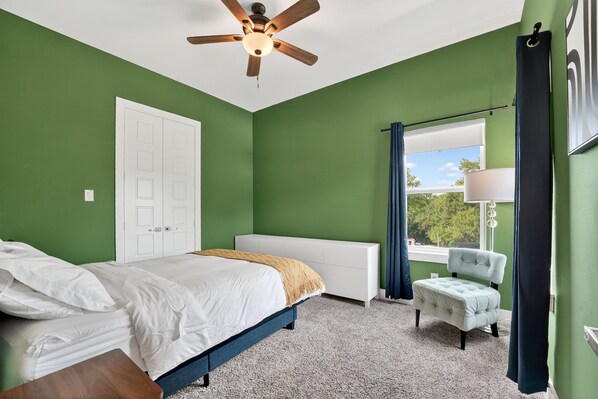 A bedroom with a bed, a chair, a window with curtains, a ceiling fan, and a door.