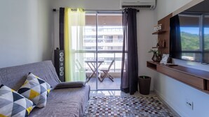 A modern living room with a grey sofa, patterned cushions, a tiled floor, a balcony with a table and chairs, and a TV on a wooden shelf. - Apto c Piscina e Quadras no Recreio (Rio de Janeiro)