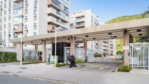A gated entrance to a residential building with a covered walkway, a motorcycle parked, and a person walking. - Apto c Piscina e Quadras no Recreio (Rio de Janeiro)