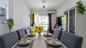 A modern dining area with a long table, grey upholstered chairs, and a vase of yellow flowers. - Apto c Piscina e Quadras no Recreio (Rio de Janeiro)